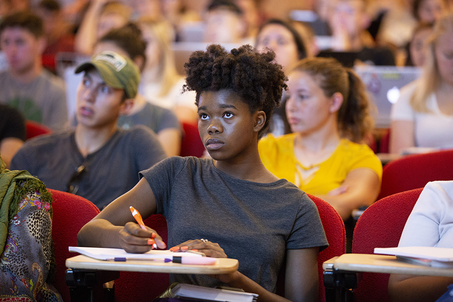 A student takes notes in the front row of a large lecture class.